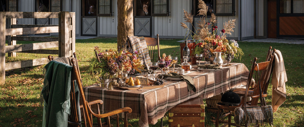 An outdoor dining table setup in a barn yard, featuring a brown and beige plaid tablecloth, wooden chairs, floral centerpieces, and cozy blankets draped over the chairs. Horses look on from barn doors. Weston Table.