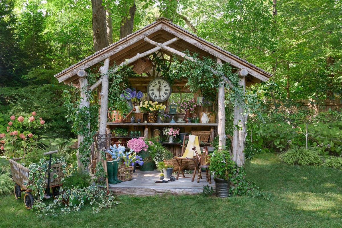 Potting shed surrounded by trees, decorated with flowers, baskets, a vintage clock, copper watering cans, and other decor.
