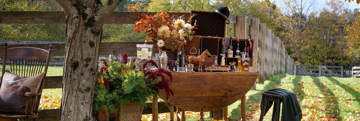 Equestrian themed outdoor bar setup with pewter and crystal barware, vintage round leaf table, stirrup cups, leather equestrian boots, and autumn maple trees. Weston Table.