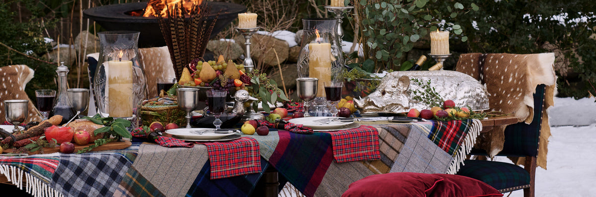 A rustic outdoor dining table set in a snowy forest, decorated with a patchwork quilt tablecloth, plaid napkins, candles, fruit, and deer-hide table runners. Weston Table.