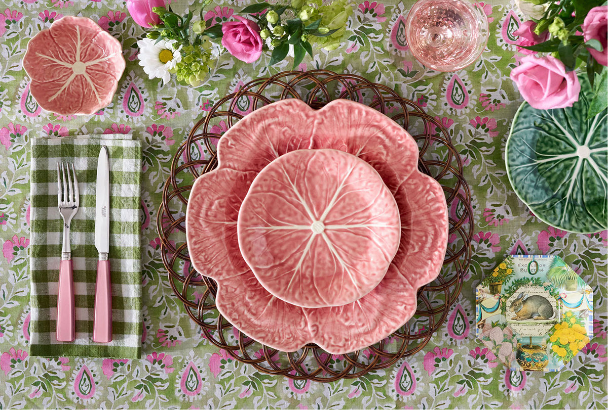 Table setting with pink Bordallo Pinheiro cabbage plates, pink flatware, and a decorative tablecloth.