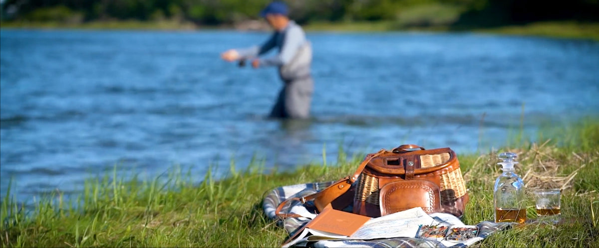 A creel and matching leather accessories are arranged on a grassy lakeshore with a blurred fly-fisher standing in the blue water in the background. Weston Table.