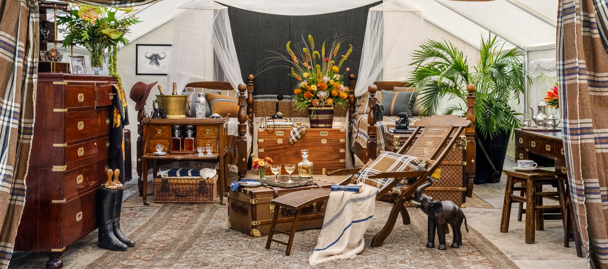 A safari tent interior with multiple wooden furniture pieces arranged under a white tent: a tall chest of drawers, side tables, a bed, a folding lounge chair, rugs, plants, and decor. Weston Table.