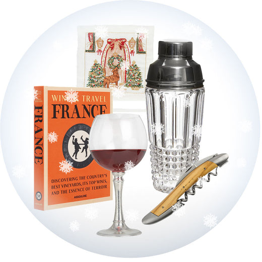 A curated gift collage featuring a wine glass with red wine, a clear textured cocktail shaker with a black lid, a wooden-handled corkscrew, a boxed travel book titled 'Wine Travel France,' and a small holiday card, arranged on a white background with a pale blue circular backdrop and faint snowflake graphics. Weston Table.