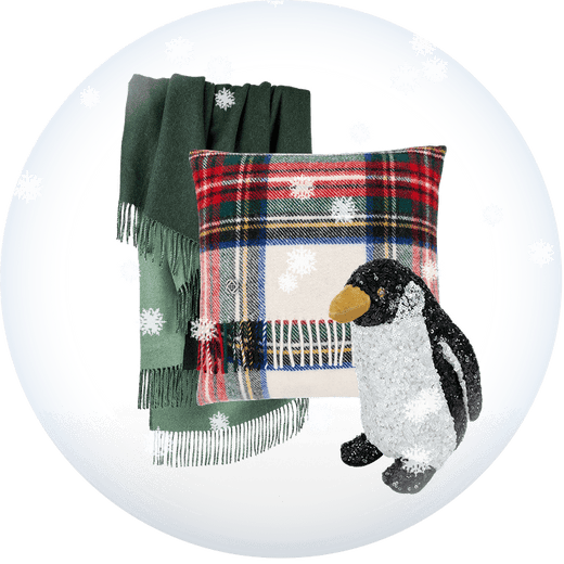 A holiday-themed product collection featuring a dark green fringed throw blanket, a red and green plaid pillow with snowflake patterns, and a plush penguin toy, all arranged inside a white circular frame with light snowflakes. Weston Table.
