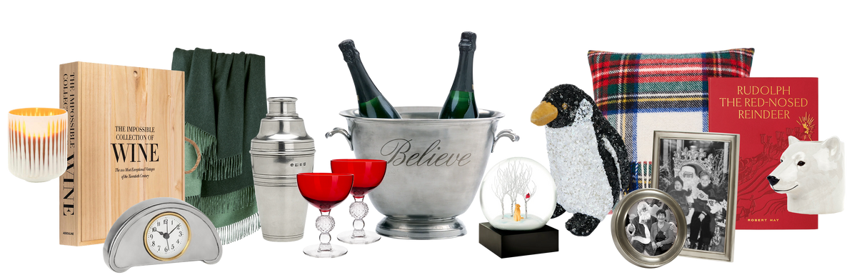 A curated collection of holiday gifts arranged on a white background, including a pewter champagne bucket engraved with the word 'Believe' with champagne bottles, red stemmed glasses, a penguin plush, a plaid throw pillow, a pewter alarm clock, a candle, a wine-themed book, a green scarf, a red book, and a black-and-white photo frame set. Weston Table.