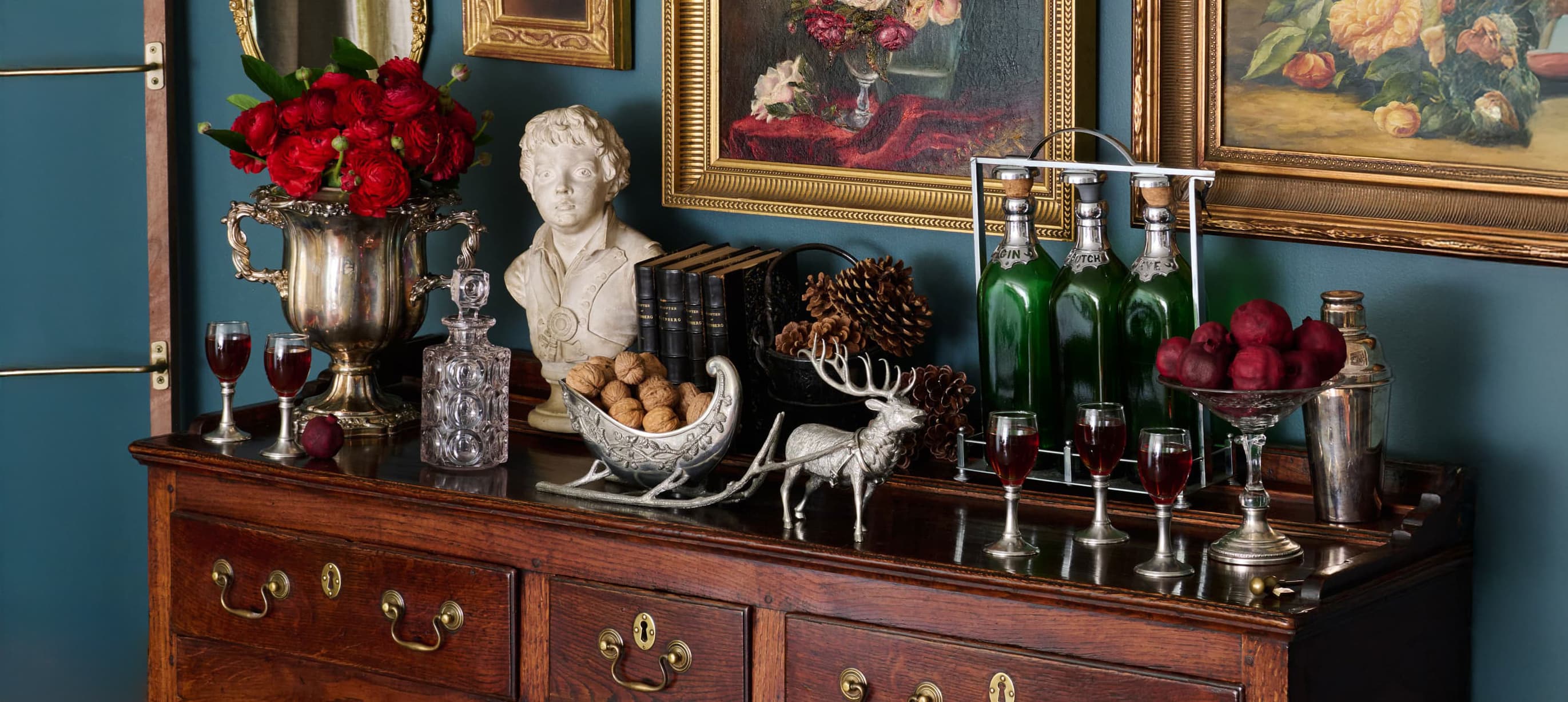 Antique sideboard with MATCH pewter wine glasses, bottles, a pewter reindeer and sleigh, and a bust against a blue wall with framed paintings.