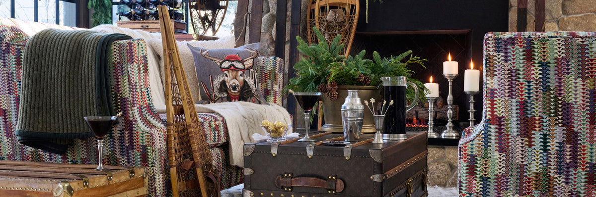 A cozy ski lodge living room scene with patterned armchairs, a rustic trunk coffee table, candles, and a fireplace in the background. Weston Table.