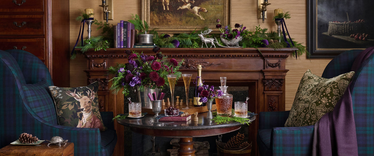 A cozy, rustic living room scene featuring a dark wood fireplace mantel decorated with greenery and purple flowers, flanked by two deep blue plaid armchairs with deer-print and purple throw pillows. A round wooden coffee table in the center holds wine glasses, decanters, and more floral arrangements. Weston Table.