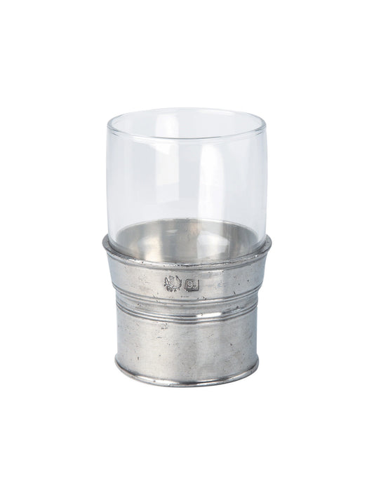 A clear glass drinking cup with a pewter sleeve, displayed on a table with a vase of flowers in the background.