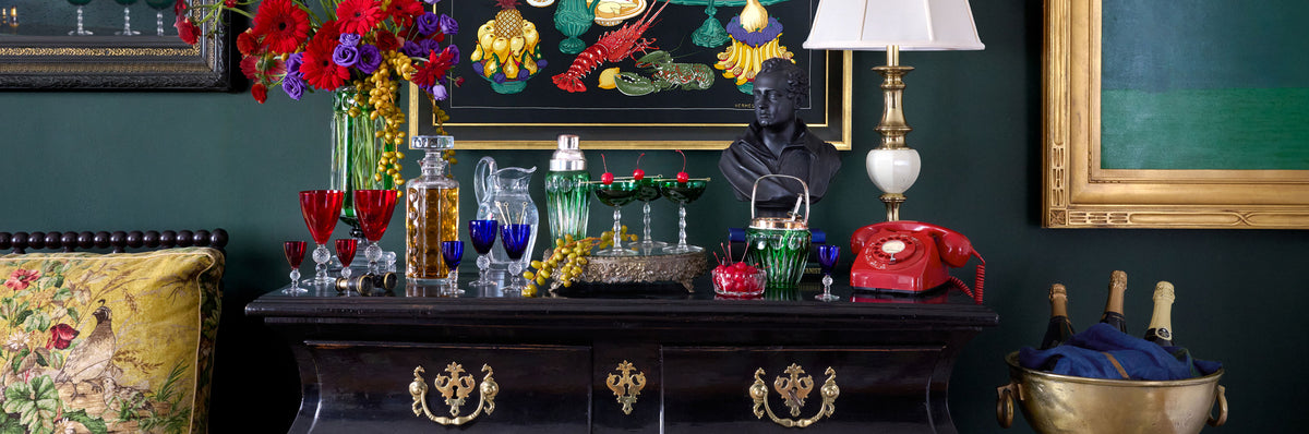 A dark wood console table in a richly decorated room, styled with a white table lamp, a red rotary telephone, a bust sculpture, colorful glassware, and a vase of vibrant flowers against a dark green wall. Weston Table.
