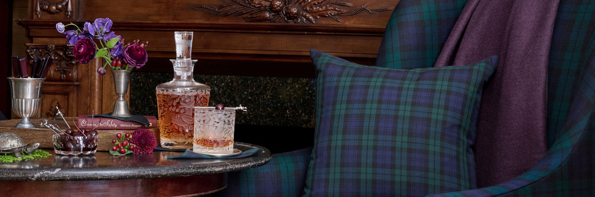 A styled interior scene with a plaid throw pillow in green and blue tartan, a matching purple throw blanket draped over an armchair, a crystal decanter and tumbler on a table, flowers in a pewter jigger, and a dark wood fireplace in the background. Weston Table.