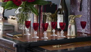  Red cordial glasses on a desk with vintage brass stirrup cups in the shape of rabbits and books and a vase of red roses. 