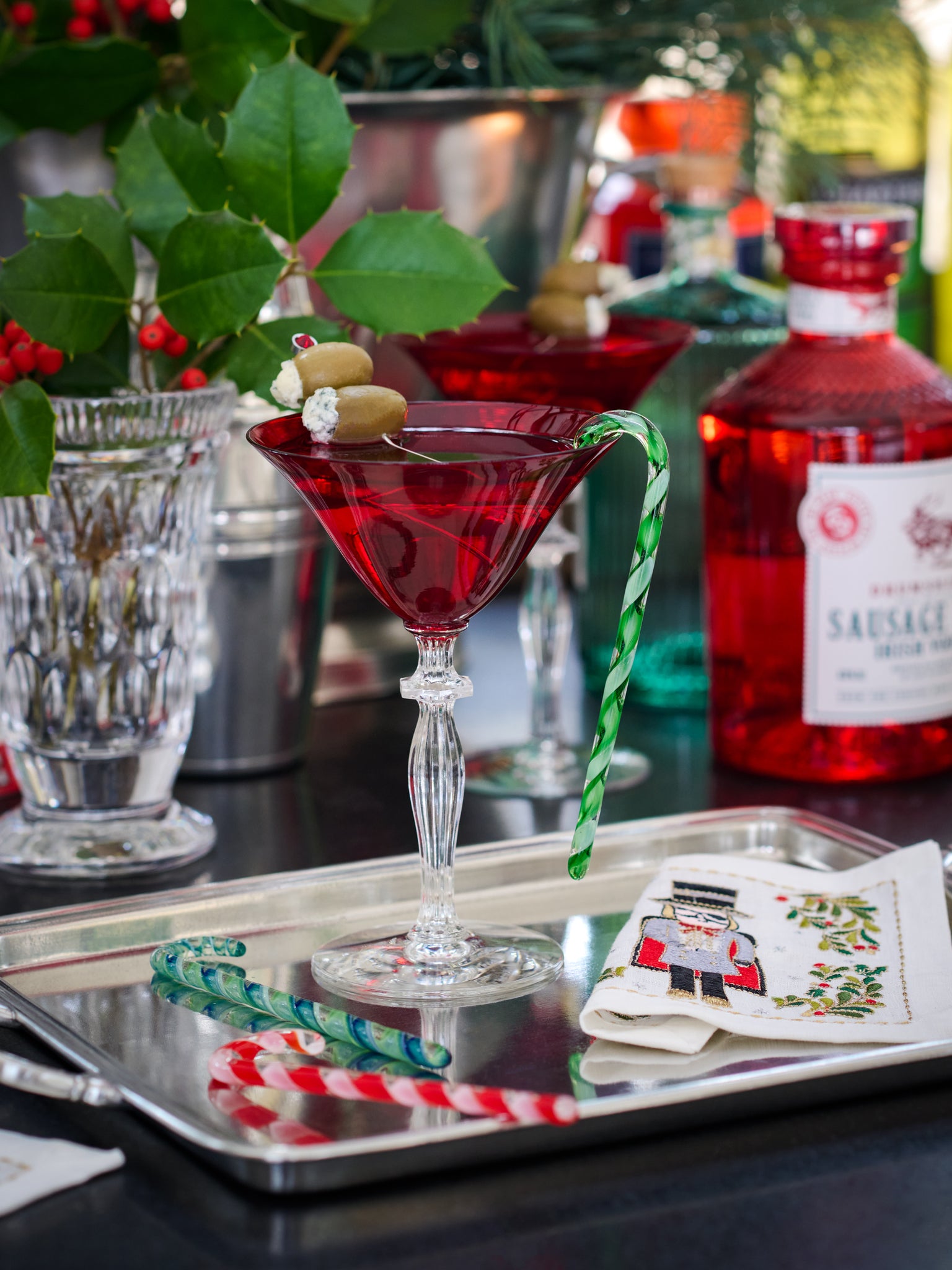 Red cocktail in a martini glass with candy canes on a tray, surrounded by festive decor.