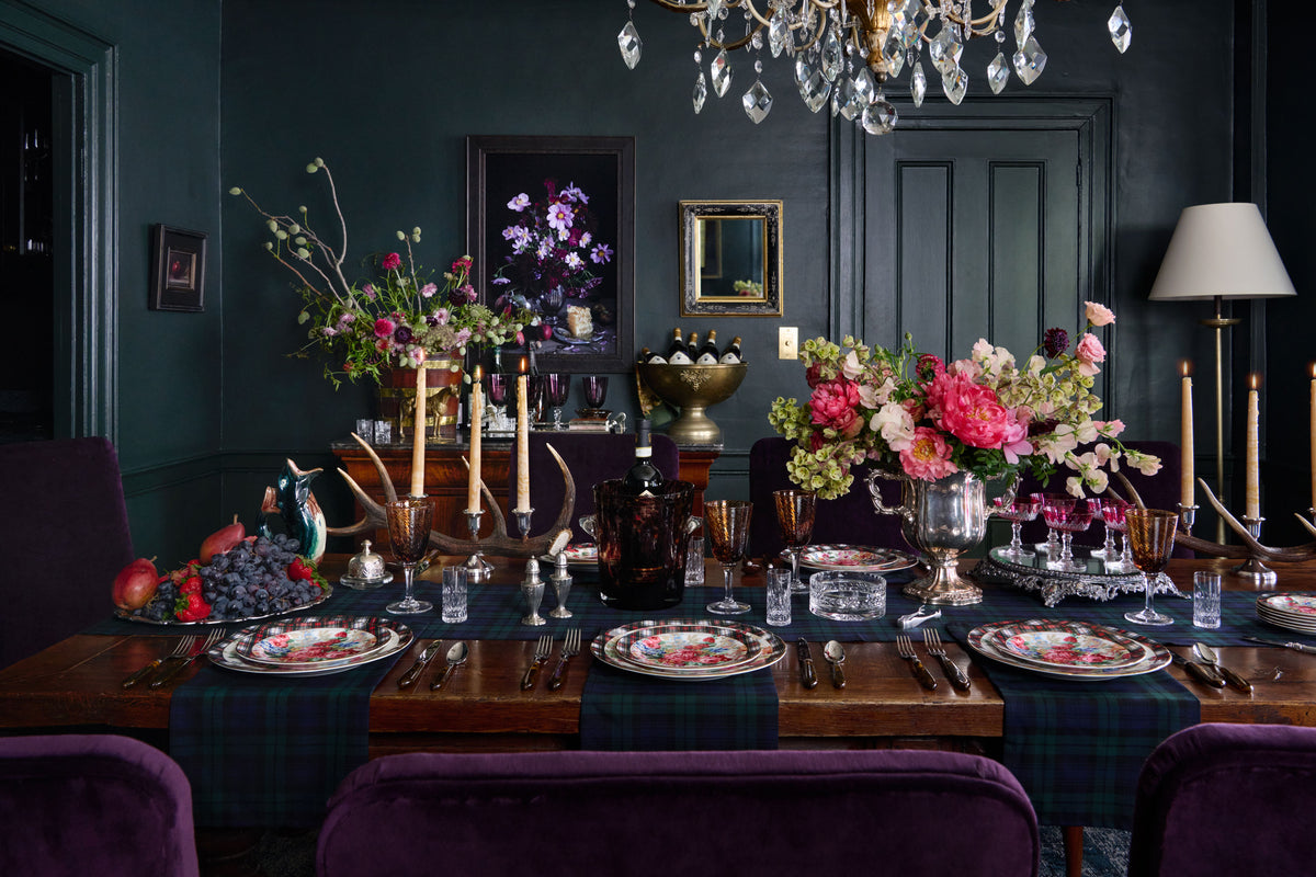 A dark, elegant dining room table set for a formal dinner, featuring deep purple velvet chairs, a green plaid table runner, floral centerpieces, crystal glassware, and ornate silver candlesticks against dark green paneled walls. Weston Table.
